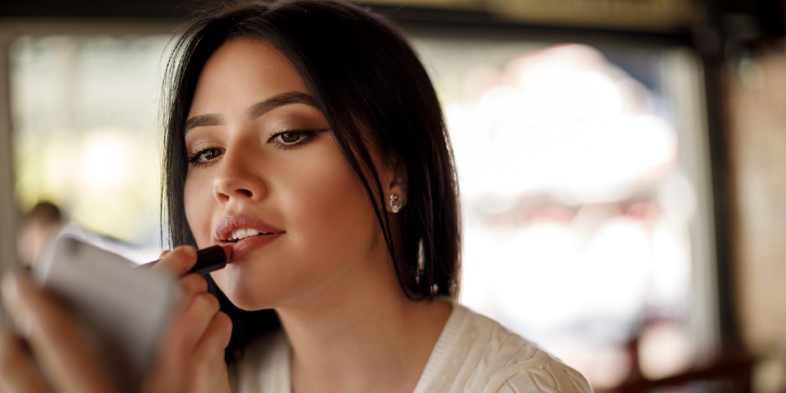 applying-matte-lipstick the-independence Young woman applying lipstick at a cafe