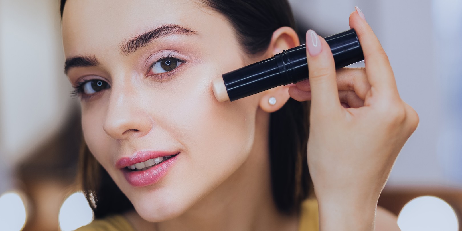 Charming woman using concealer stick while putting makeup on