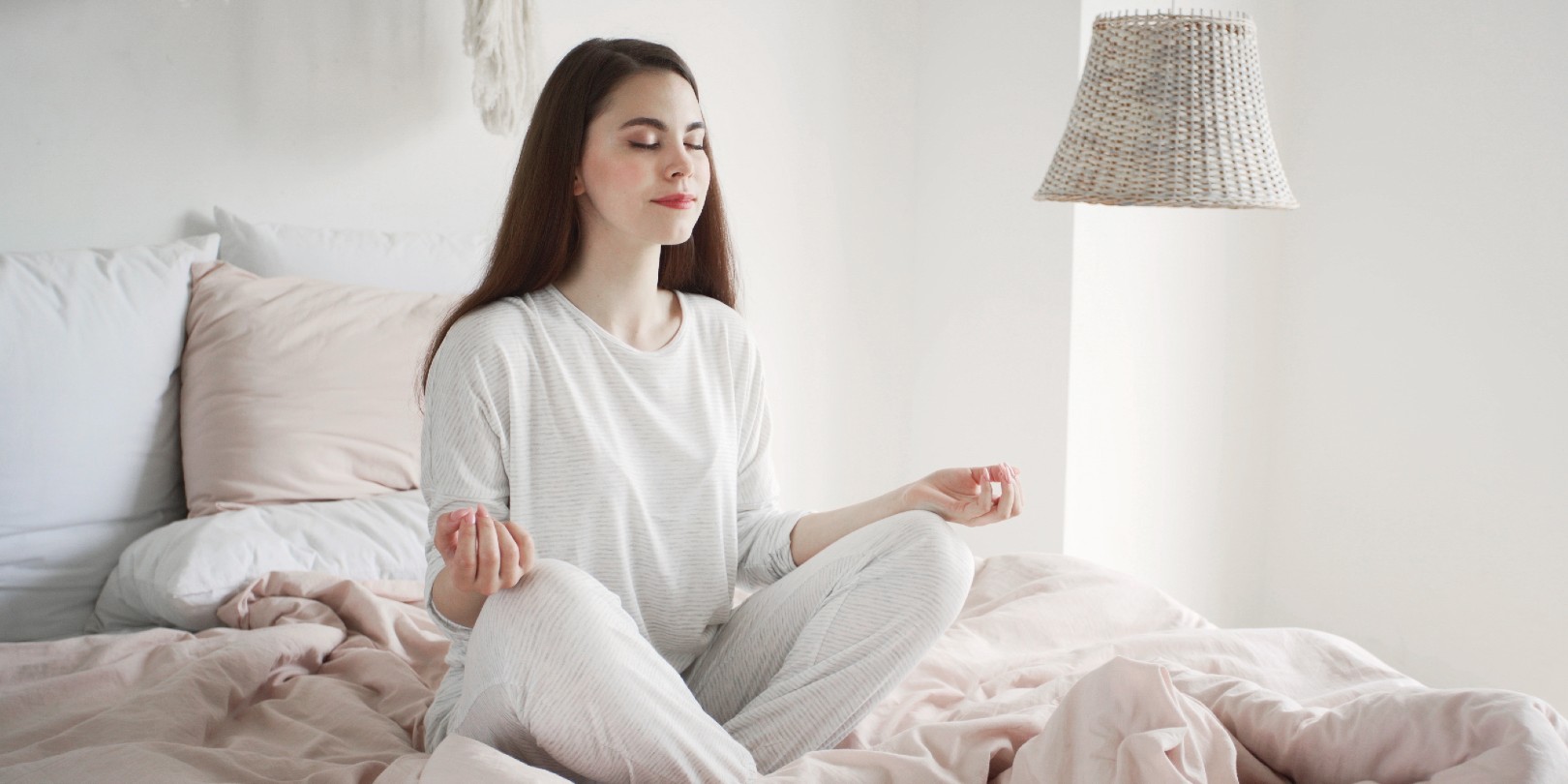 Calm and dreamy young woman in pajamas sitting at meditation pose on bed in cozy bedroom