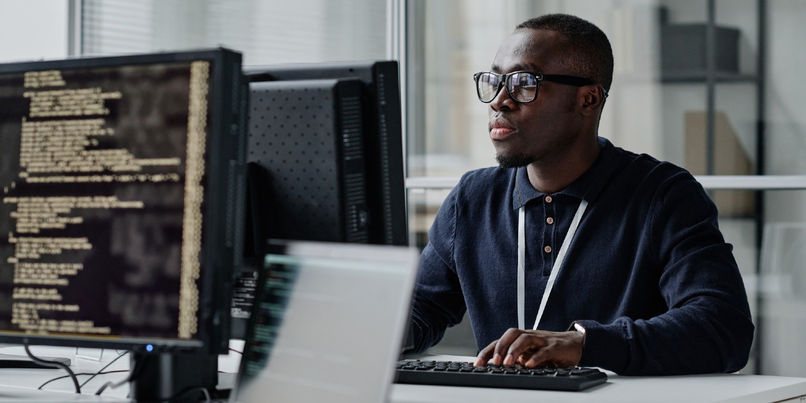 African American young developer in eyeglasses concentrating on his online work on computer sitting at workplace