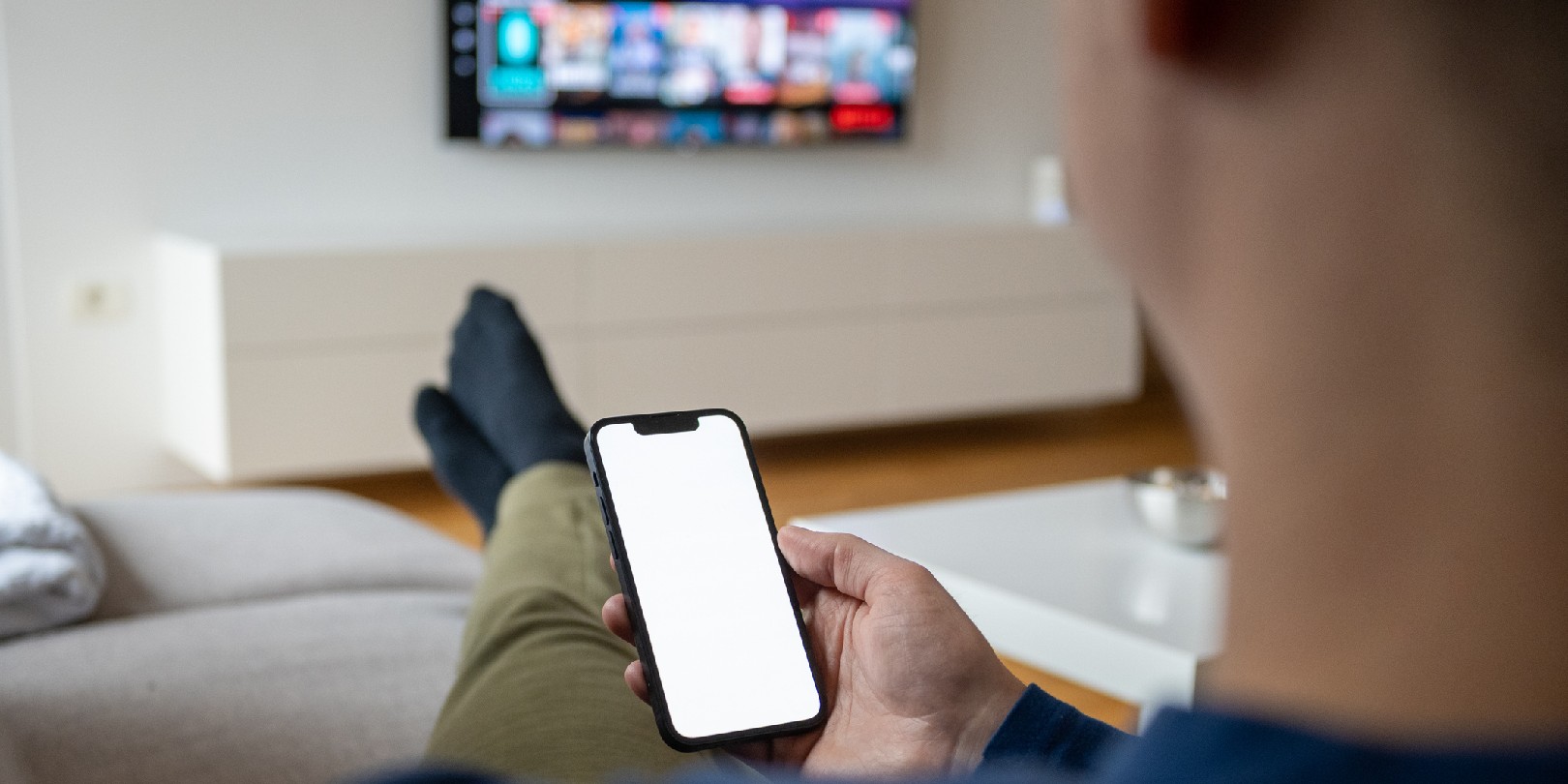 Over the shoulder view of a caucasian man holding a smart phone with a white screen while watching TV.
