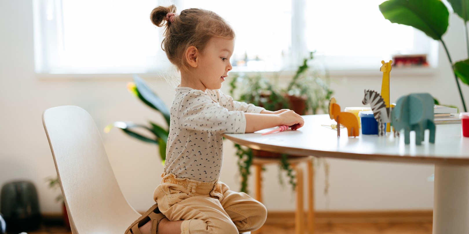 Cute little girl is crouching on the chair playing with paper animals that are on the table.