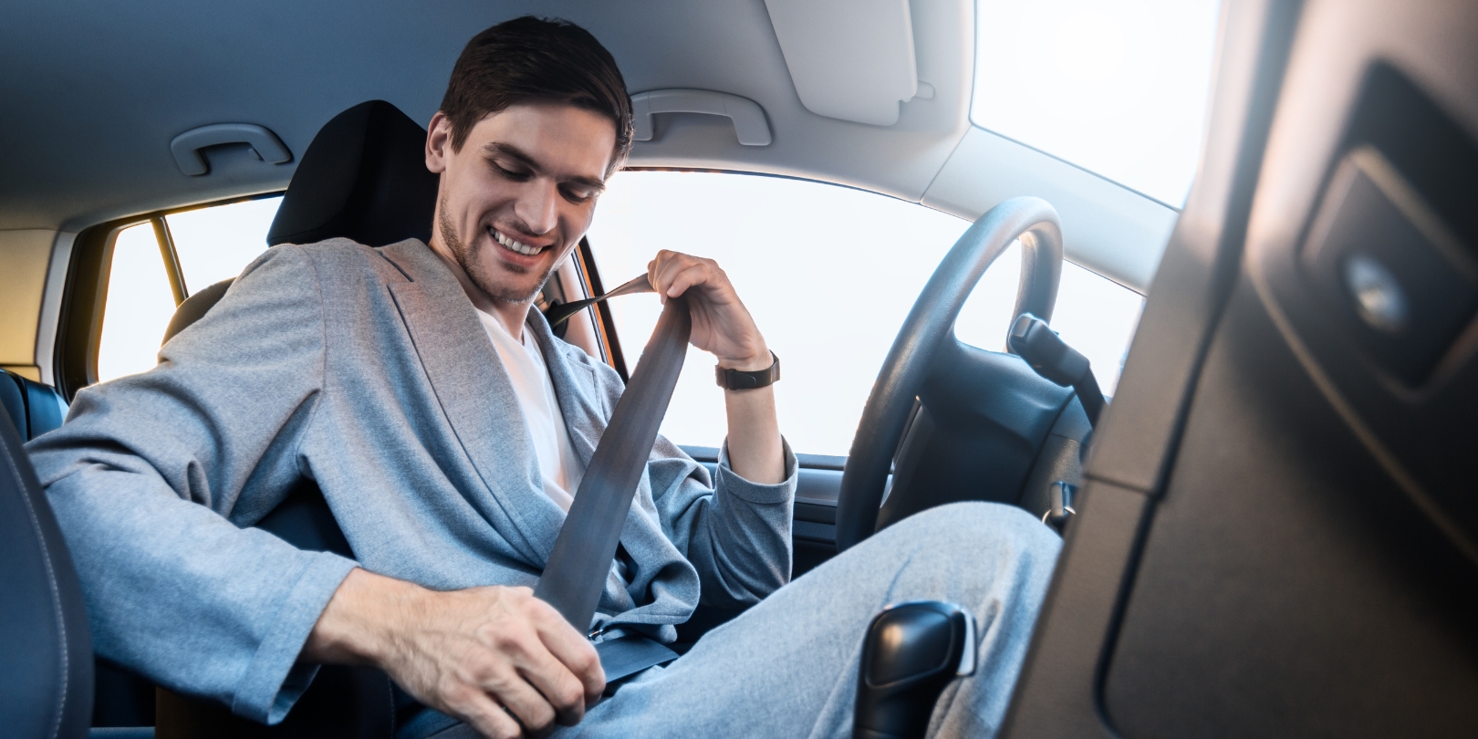 Closeup driver is fastening his seat belt. Young businessman in grey suit is riding behind steering wheel of car. Man is following rules of road. Concept of route safety in modern city.