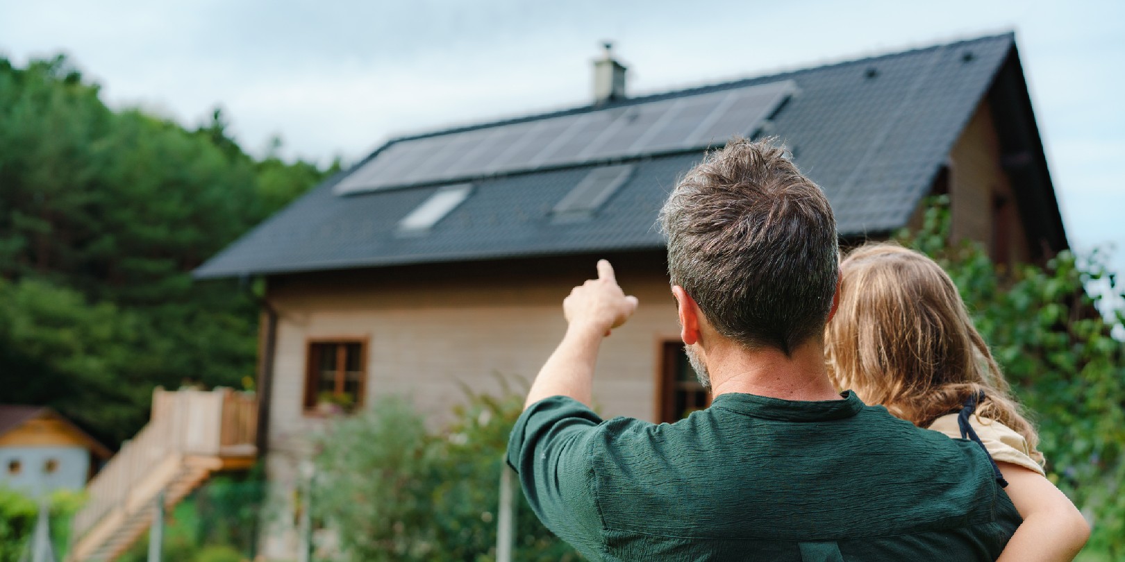 eco-houses the-independence Rear view of dad holding her little girl in arms and showing at their house with solar panels.Alternative energy, saving resources and sustainable lifestyle concept.