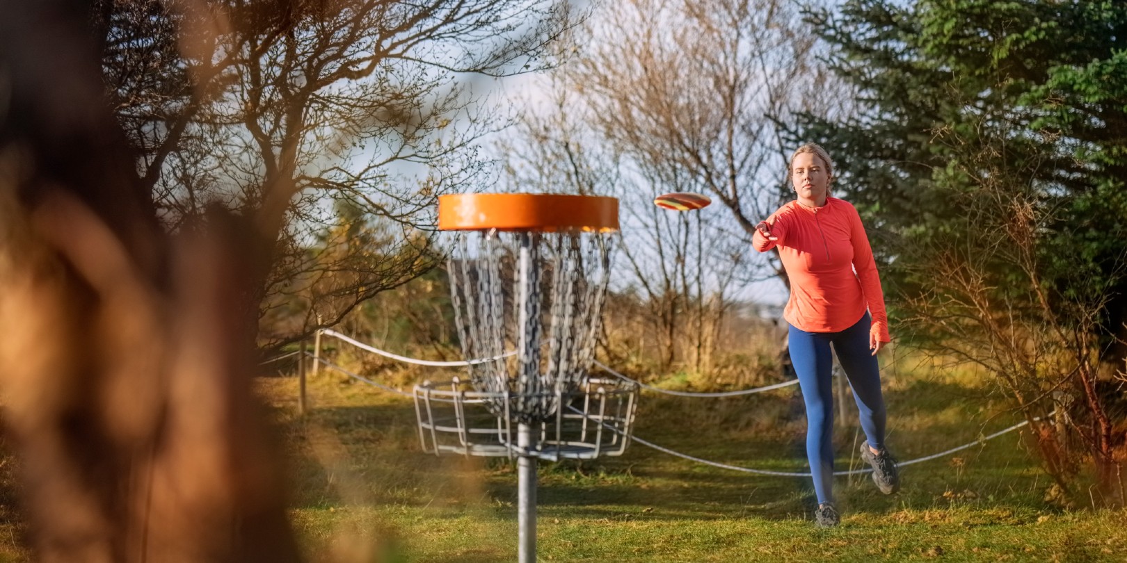 Woman tossing a disc into the basket goal. Caucasian woman playing disc golf game outdoors in a park.